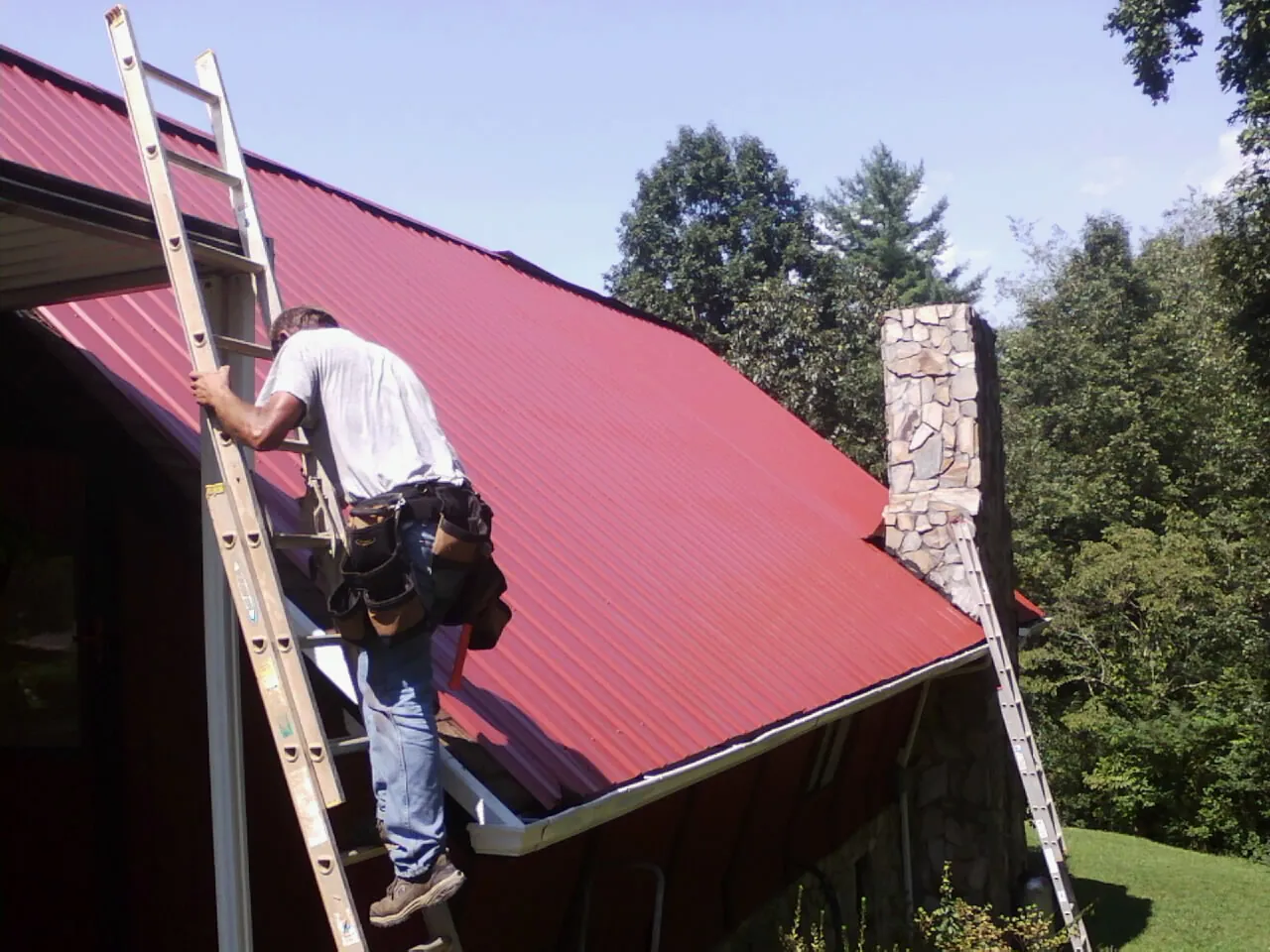 Roofer on a ladder beside a red metal roof and stone chimney — Soffit & Fascia Repair by Roofing Brothers in Harrisburg